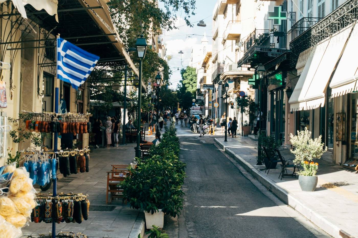 A shaded street in central Athens with shopfronts, trees and a Greek flag over the pavement