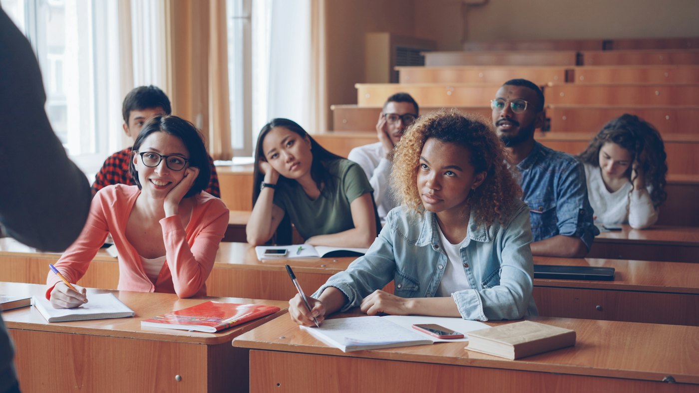 Students taking notes in a university lecture room while a lecturer speaks at the front